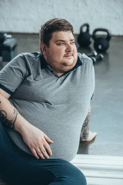 Overweight tattooed man looking away while sitting on fitness mat at sports center — Stock Photo