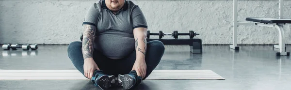 Panoramic shot of overweight man sitting on fitness mat and stretching legs at sports center — Stock Photo