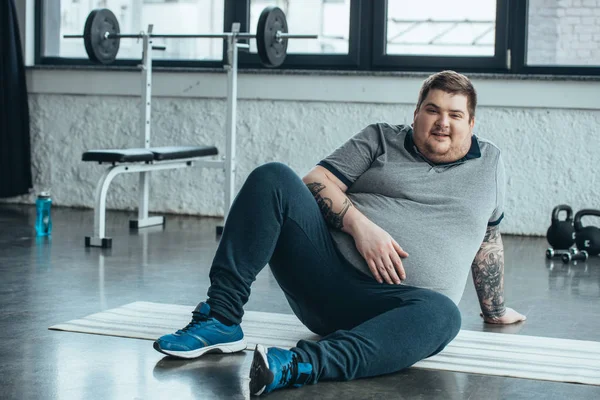 Hombre tatuado con sobrepeso mirando a la cámara y sentado en la colchoneta de fitness en el centro deportivo - foto de stock