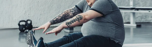 Panoramic shot of overweight tattooed man sitting and stretching at gym — Stock Photo