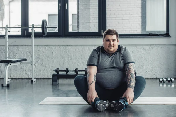 Sovrappeso tatuato uomo seduto e guardando fotocamera durante l'esercizio di stretching in palestra — Foto stock