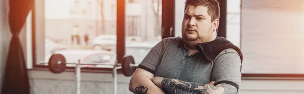 Sobrepeso hombre tatuado con toalla y brazos cruzados mirando a la cámara en el gimnasio con sol - foto de stock