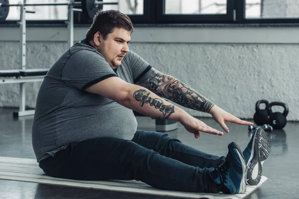 Overweight tattooed man sitting on fitness mat and stretching at gym — Stock Photo