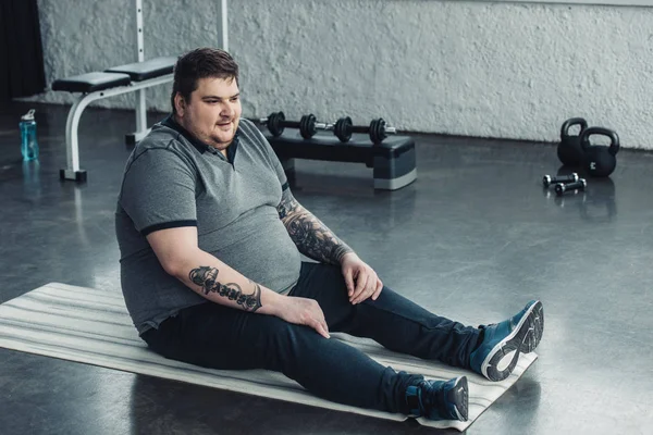 Overweight tattooed man in grey t-shirt sitting on fitness mat at sports center — Stock Photo