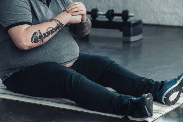 Обрезанный вид толстого татуированного мужчины, сидящего на тренажерном коврике и вытягивающего пальцы в спортзале — стоковое фото