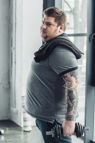 Obese tattooed man looking at camera while exercising with dumbbell at sports center — Stock Photo