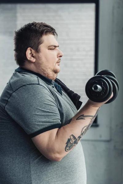 Vista lateral do homem tatuado Obese exercitando com halteres no centro de esportes — Fotografia de Stock