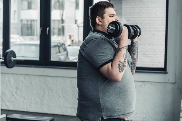 Hombre tatuado con sobrepeso haciendo ejercicio con pesas en el gimnasio con espacio para copiar - foto de stock