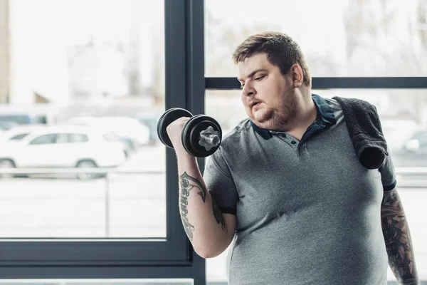 Overweight tattooed man with towel exercising with dumbbell at gym with copy space — Stock Photo