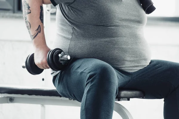 Vista recortada del hombre tatuado con sobrepeso sentado y entrenando con mancuerna en el gimnasio - foto de stock