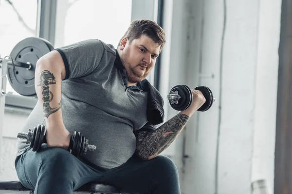 Ожирний татуйований чоловік займається з гантелями і дивиться на камеру в спортивному центрі — стокове фото
