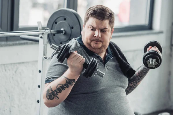 Ожирний татуйований чоловік займається з гантелями в спортивному центрі — стокове фото