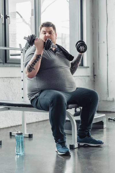 Overweight tattooed man sitting and exercising with dumbbells at sports center — Stock Photo