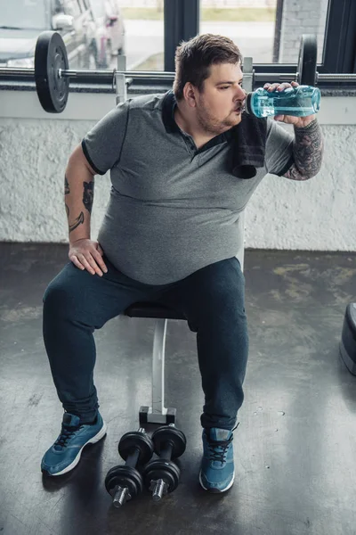 Overweight tattooed man drinking water from sport bottle after training with dumbbells at sports center — Stock Photo