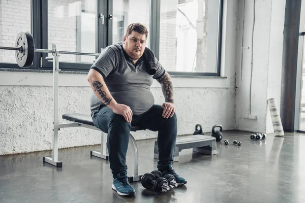 Stanco uomo obeso seduto sulla panchina e guardando fotocamera dopo l'allenamento con manubri in palestra — Foto stock