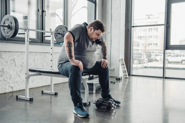 Müder übergewichtiger Mann sitzt nach Training mit Kurzhanteln im Fitnessstudio auf Bank — Stockfoto