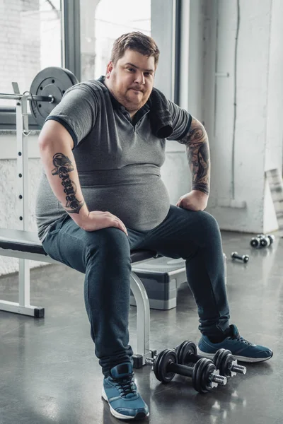 Cansado Hombre obeso sentado en el banco y mirando a la cámara después de hacer ejercicio con pesas en el gimnasio - foto de stock