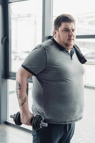 Избыточный вес татуированный мужчина в серой футболке с полотенцем упражнения с гантелями в спортивном центре — стоковое фото