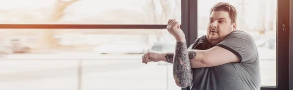 Panoramic shot of overweight tattooed man stretching arms at gym with copy space — Stock Photo