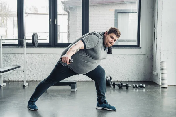 Übergewichtiger, tätowierter Mann im grauen T-Shirt mit Handtuch trainiert mit Hantel im Sportzentrum — Stockfoto