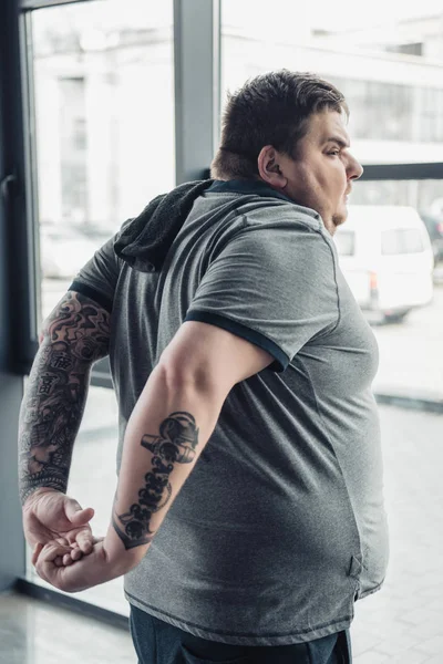 Sobrepeso tatuado homem em branco t-shirt alongamento braços no esportes centro — Fotografia de Stock