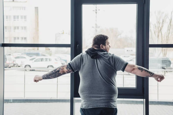 Back view of overweight tattooed man in grey t-shirt stretching at gym — Stock Photo