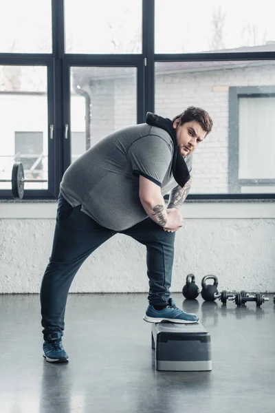 Übergewichtiger tätowierter Mann mit Handtuch blickt beim Training auf Trittbrett im Sportzentrum in die Kamera — Stockfoto