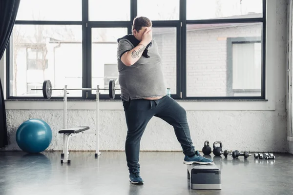 Übergewichtiger Mann mit Handtuch bedeckt Gesicht mit Hand beim Training auf Trittbrett im Sportzentrum — Stockfoto