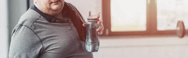 Panoramic shot of overweight man holding sport bottle at gym with sunlight and copy space — Stock Photo