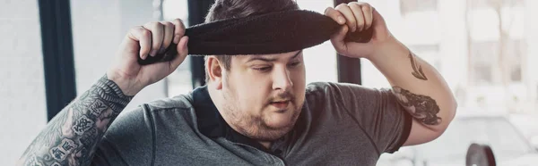 Panoramic shot of tired overweight tattooed man wiping face with towel at sports center — Stock Photo