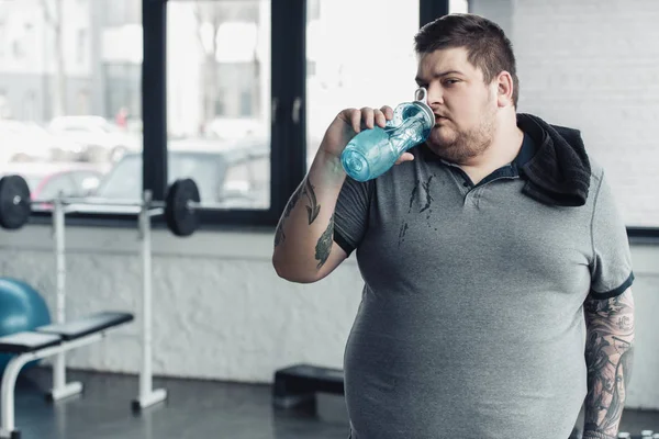Sobrepeso hombre tatuado con toalla agua potable de la botella de deporte en el gimnasio - foto de stock