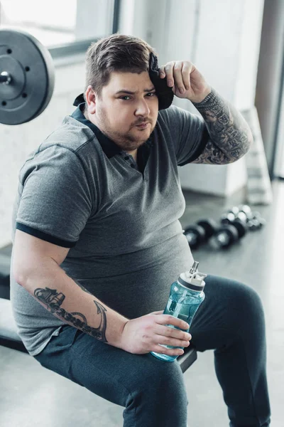 Sobrepeso tatuado homem segurando esporte garrafa, olhando para câmera e limpando rosto com toalha no ginásio — Fotografia de Stock