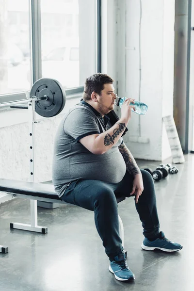 Overweight tattooed man sitting on bench and drinking water from sport bottle at gym — Stock Photo