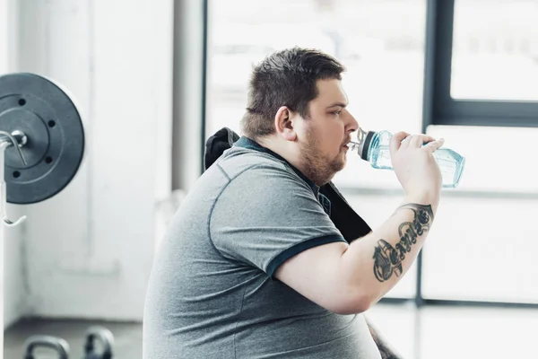 Vista lateral del hombre tatuado con sobrepeso bebiendo agua de la botella de deporte en el gimnasio - foto de stock