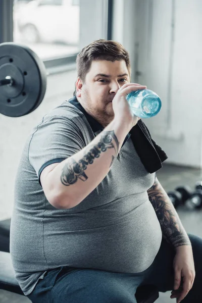 Hombre tatuado con sobrepeso cansado bebiendo agua de la botella de deporte en el gimnasio - foto de stock