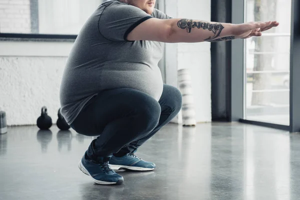 Abgeschnittene Ansicht eines übergewichtigen tätowierten Mannes bei Kniebeugen im Sportzentrum — Stockfoto