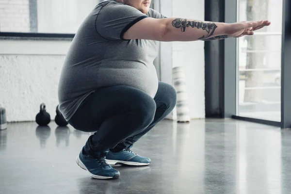 Vista recortada del hombre tatuado con sobrepeso haciendo sentadillas en el centro deportivo - foto de stock