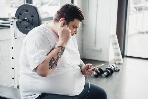 Übergewichtiger Mann mit Kopfhörer sitzt auf Bank und benutzt Smartphone im Fitnessstudio — Stockfoto