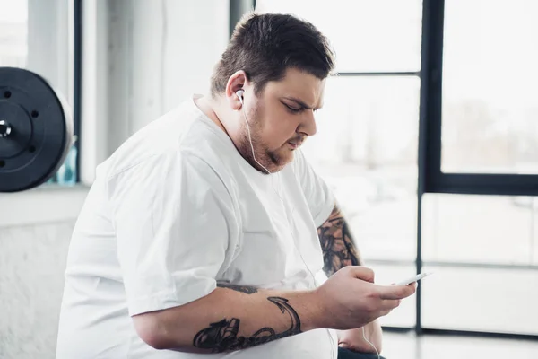 Overweight man in earphones sitting and using smartphone at gym — Stock Photo