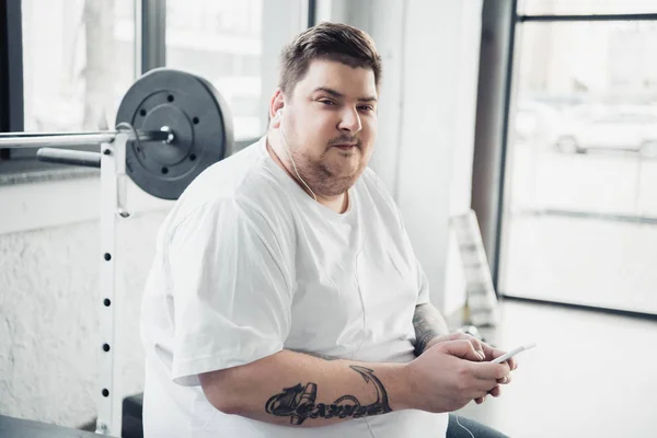 Übergewichtiger Mann mit Kopfhörer sitzt auf Bank, blickt in Kamera und benutzt Smartphone im Fitnessstudio — Stockfoto