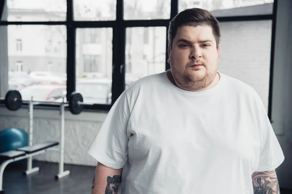 Sobrepeso tatuado homem em branco t-shirt olhando para câmera no centro de esportes com espaço de cópia — Fotografia de Stock