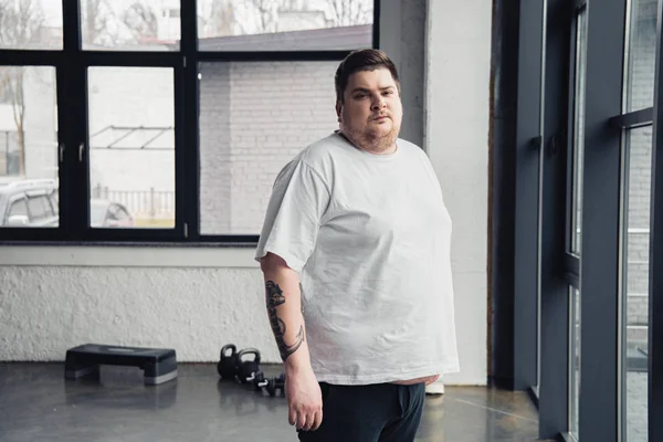 Надмірна вага татуйованого чоловіка в білій футболці, дивлячись на камеру в спортивному центрі з копіювальним простором — стокове фото