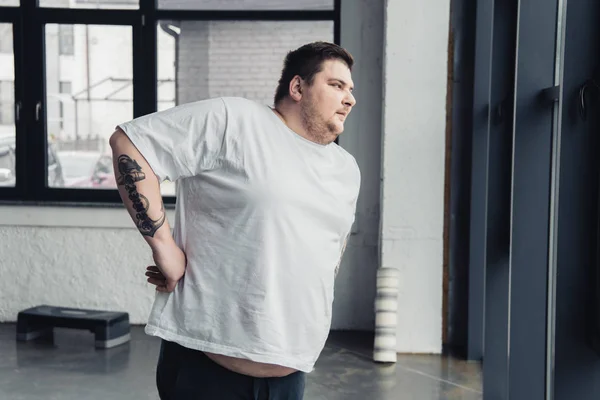Obese tattooed man in white t-shirt doing stretching exercise at sports center — Stock Photo