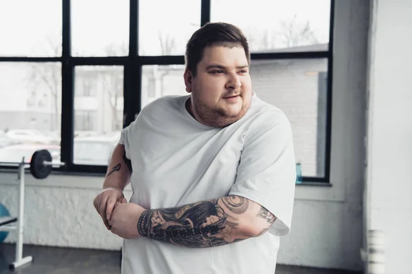 Overweight tattooed man in white t-shirt stretching arms at gym — Stock Photo