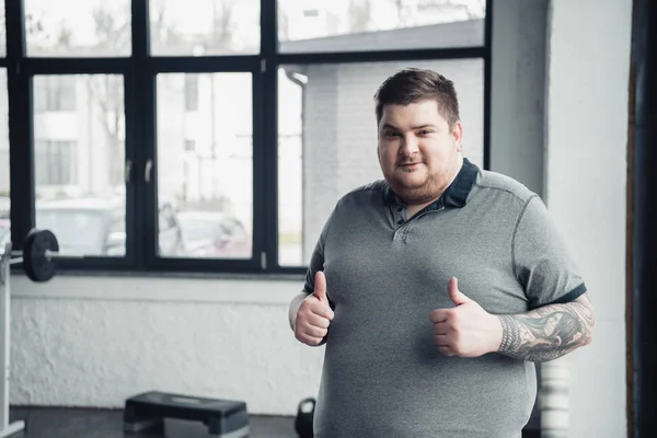 Sovrappeso tatuato uomo guardando fotocamera e mostrando pollici in palestra con spazio copia — Foto stock