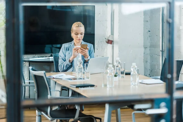 Selective focus of attractive blonde businesswoman sitting in conference room and using smartphone — Stock Photo