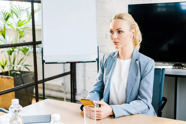 Привлекательная блондинка бизнес-тренер сидит в конференц-зале и держит смартфон — стоковое фото
