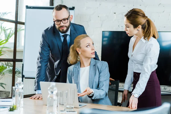 Atractiva mujer de negocios gesto mientras está sentado cerca de la computadora portátil y mirando a su compañero de trabajo - foto de stock