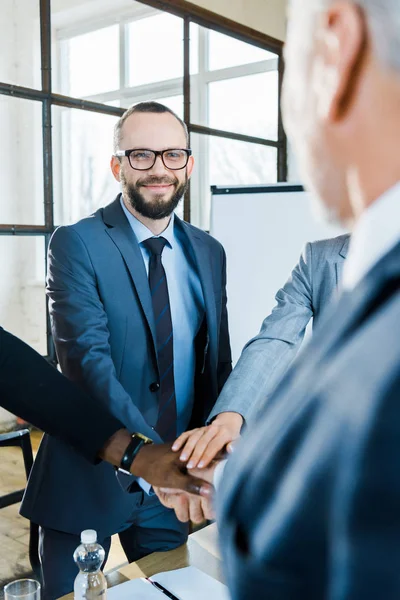 Hombre barbudo feliz poniendo manos juntas con compañeros de trabajo multiculturales en la sala de conferencias - foto de stock