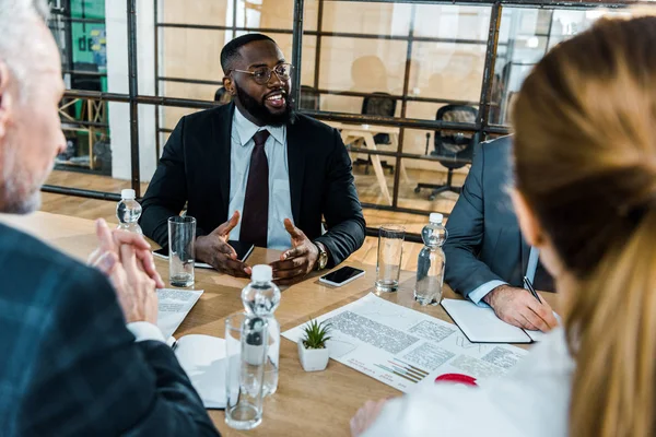 Selective focus of cheerful african american man in eye glasses sitting in conference room with coworkers — Stock Photo